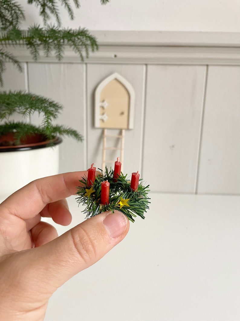 May include: A miniature Christmas wreath with five red candles and small yellow star accents. The wreath is held in a hand, with a miniature door and ladder in the background. The wreath is made of green faux pine needles.