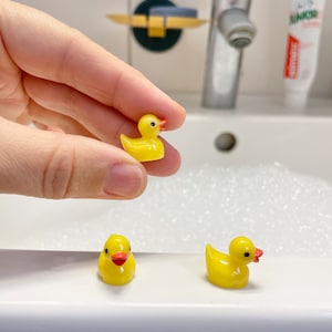 Könnte beinhalten: Drei kleine gelbe Quietscheenten mit orangefarbenen Schnäbeln und schwarzen Augen. Eine Ente wird von einer Hand gehalten, die anderen beiden sitzen auf einer weißen Oberfläche. Die Enten befinden sich in einem Badezimmer, in der Nähe eines Wasserhahns und Zahnpasta.