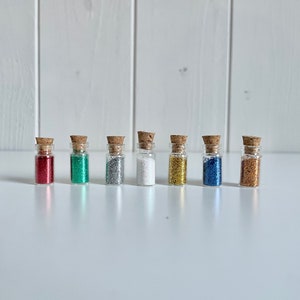 Könnte beinhalten: Sieben kleine Glasflaschen mit Korken, jede gefüllt mit einer anderen Farbe Glitzer. Die Farben sind Rot, Grün, Silber, Weiß, Gold, Blau und Bronze. Die Flaschen sind in einer Reihe auf einer weißen Oberfläche angeordnet.