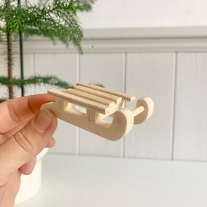 Könnte beinhalten: Ein kleiner Holzschlitten mit natürlicher Oberfläche. Der Schlitten hat eine Lattenplattform und abgerundete Kufen. Das Design ist schlicht und rustikal, ideal für Weihnachtsdekorationen oder Miniaturpräsentationen. Der Schlitten wird in einer Hand gehalten.