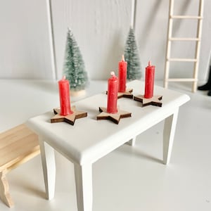 Könnte beinhalten: Ein kleiner weißer Tisch mit vier roten Kerzen auf sternförmigen Kerzenhaltern aus Holz. Die Kerzen sind angezündet. Im Hintergrund stehen zwei kleine grüne Weihnachtsbäume. Der Tisch ist 30 cm lang.