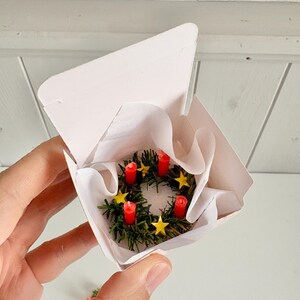 May include: Miniature Advent wreath with four red candles and yellow star decorations, nestled in a white paper-lined box. The wreath is made of green faux evergreen branches. A festive holiday decoration.