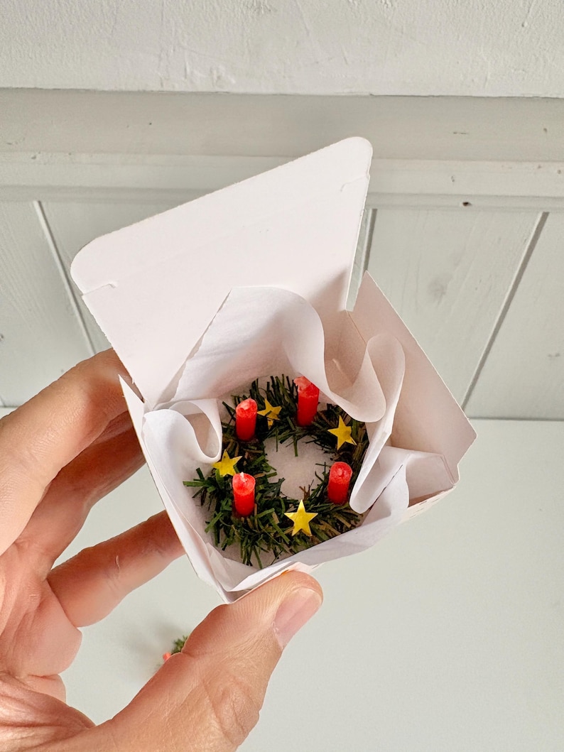 May include: Miniature Advent wreath with four red candles and yellow star decorations, nestled in a white paper-lined box. The wreath is made of green faux evergreen branches. A festive holiday decoration.