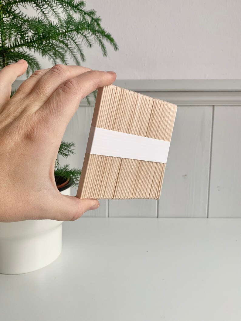 May include: A bundle of natural wood craft sticks, approximately 50 sticks, stacked and held in a hand. The sticks are bundled together with a white label.