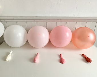 May include: A set of four balloons in white, pink, and rose gold. The balloons are arranged in a row on a white surface. There are also three deflated balloons in white, pink, and rose gold.
