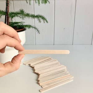 May include: A pile of natural wooden craft sticks, with one stick held up by a hand. The sticks are about 6 inches long and 1/2 inch wide.