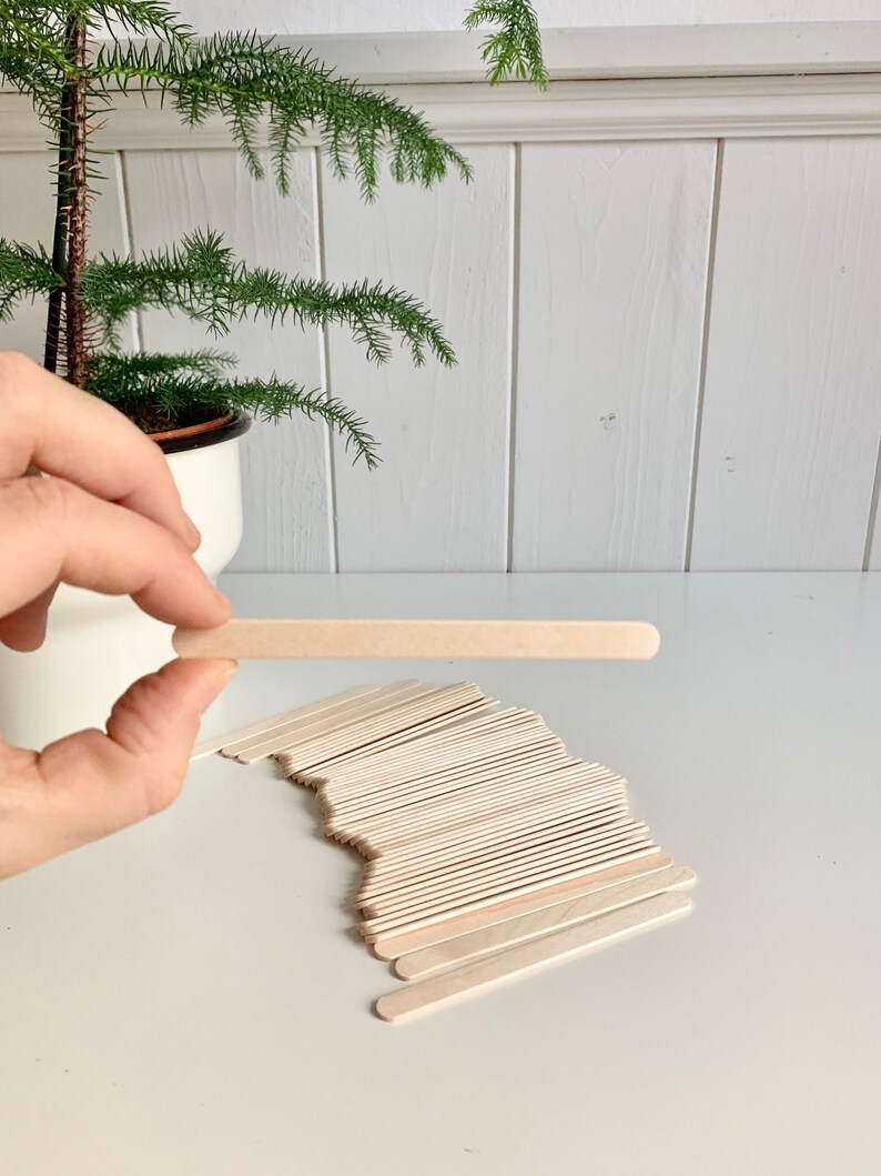 May include: A pile of natural wooden craft sticks, with one stick held up by a hand. The sticks are about 6 inches long and 1/2 inch wide.