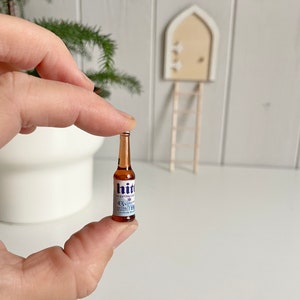 May include: A miniature brown glass bottle of "hit" beer with a white and blue label. The bottle is held between fingers. The label includes the text "EXTRA COLD" and "4.3% vol".