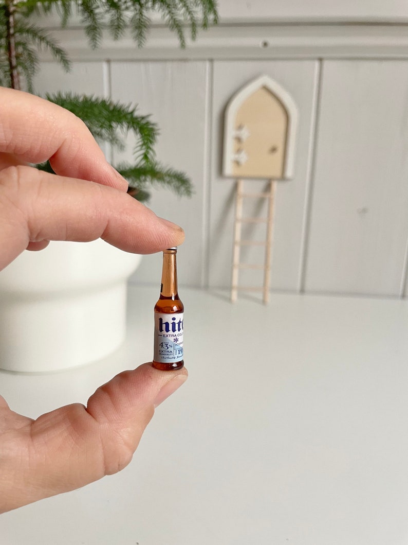 May include: A miniature brown glass bottle of "hit" beer with a white and blue label. The bottle is held between fingers. The label includes the text "EXTRA COLD" and "4.3% vol".