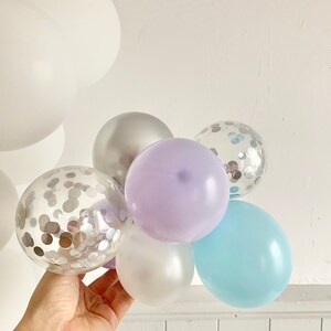 May include: A cluster of balloons in various colors, including silver, lavender, light blue, and clear with silver confetti. The balloons are tied together and held in a hand.
