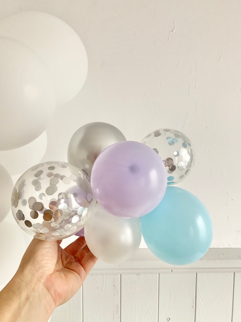 May include: A cluster of balloons in various colors, including silver, lavender, light blue, and clear with silver confetti. The balloons are tied together and held in a hand.