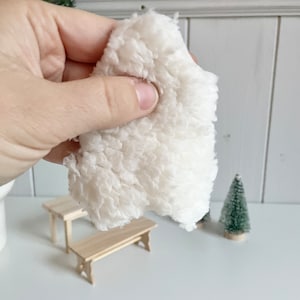 Könnte beinhalten: Ein Stück flauschigen, cremefarbenen Stoffes wird von einer Hand gehalten. Im Hintergrund befinden sich Miniaturmöbel aus Holz und ein kleiner grüner Weihnachtsbaum. Der Stoff hat eine weiche, strukturierte Oberfläche.