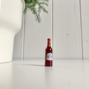 May include: A miniature brown beer bottle with a red and white label, featuring the brand name "Budweiser". The bottle is positioned on a white surface, with a white pot and green plant in the background.