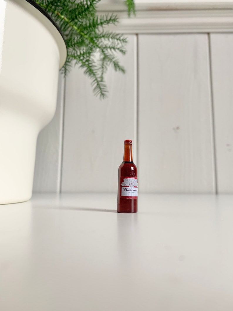 May include: A miniature brown beer bottle with a red and white label, featuring the brand name "Budweiser". The bottle is positioned on a white surface, with a white pot and green plant in the background.