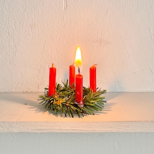 May include: A small, festive Advent wreath with four red candles, one lit with a bright yellow flame. The candles are set in a bed of green pine needles and small gold accents, creating a warm, inviting holiday scene.
