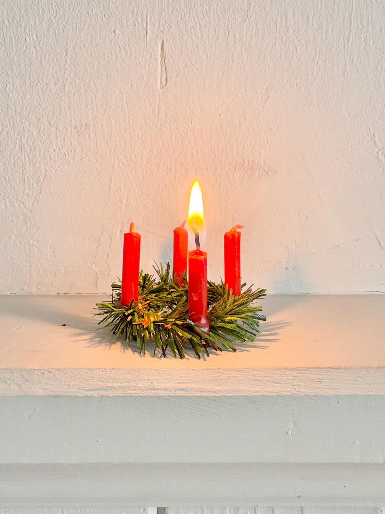 May include: A small, festive Advent wreath with four red candles, one lit with a bright yellow flame. The candles are set in a bed of green pine needles and small gold accents, creating a warm, inviting holiday scene.