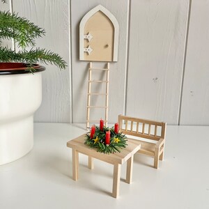 May include: A miniature wooden fairy garden scene featuring a small table with a wreath of red candles, a bench, a ladder, and a door. The door is cream-colored with star accents. A potted plant is in the background.