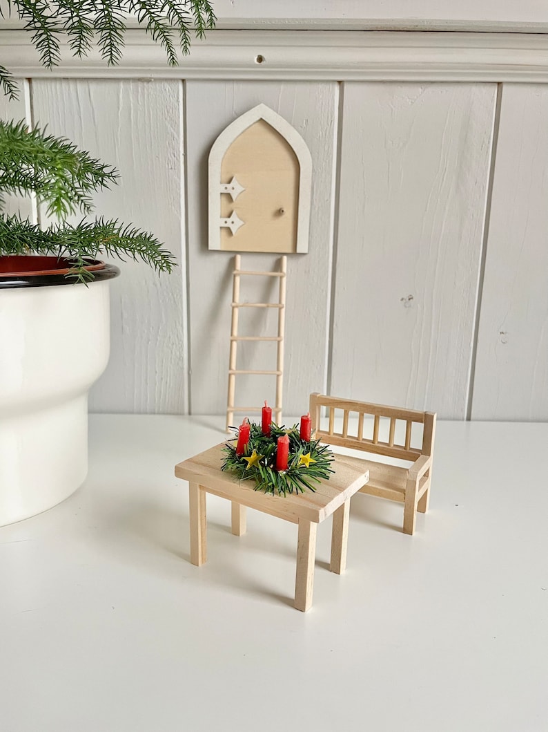 May include: A miniature wooden fairy garden scene featuring a small table with a wreath of red candles, a bench, a ladder, and a door. The door is cream-colored with star accents. A potted plant is in the background.