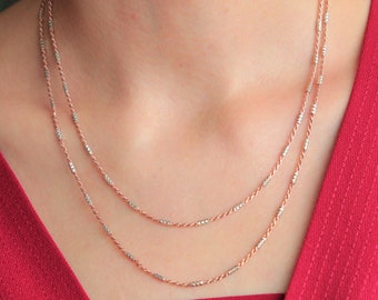 May include: A layered necklace with a rose gold chain and silver accents. The necklace is made of small, round beads and is worn by a person with a red shirt.