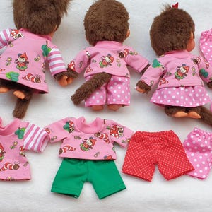 Könnte beinhalten: Drei braune Affenpuppen, die rosa Outfits mit einem skurrilen Aufdruck eines Zwergs, Pilzen und Bäumen tragen. Zusätzliche rosa und grüne Shorts und Röcke sind ebenfalls abgebildet. Die Outfits haben weiß gestreifte Ärmel.