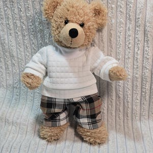 Könnte beinhalten: Ein Teddybär mit hellbraunem Fell, der einen weißen, gesteppten Pullover und karierte Shorts trägt. Die Shorts haben ein schwarz-weiß-rotes Karomuster. Der Bär hat eine schwarze Nase und steht auf einer strukturierten Oberfläche.