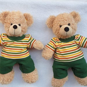 Könnte beinhalten: Zwei identische Plüschbären mit hellbraunem Fell, schwarzen Augen und schwarz gestickten Nasen. Jeder Bär trägt ein kurzärmeliges T-Shirt mit horizontalen Streifen in Grün, Gelb und Weiß sowie eine dazu passende dunkelgrüne kurze Hose.