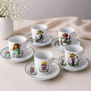 Può includere: Un set di cinque tazze e piattini in ceramica bianca, ognuno caratterizzato da un'illustrazione colorata unica di un bambino con cappello circondato da fiori. I piattini hanno un sottile bordo verde e le tazze presentano scene di giardino.