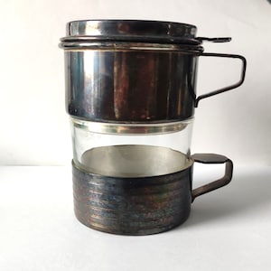 May include: A vintage Vietnamese coffee filter, featuring a stainless steel upper chamber and a clear glass carafe. The filter has a handle for easy use and a classic design. The metal has a tarnished appearance.