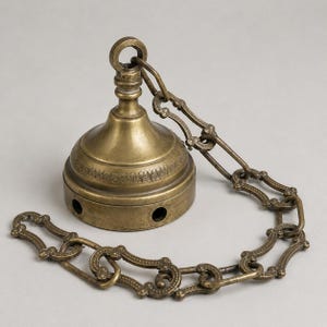 Puede incluir: Un dosel de techo de latón de estilo vintage con una cadena decorativa. El dosel tiene forma de campana con un patrón detallado alrededor de la base. Los eslabones de la cadena presentan un diseño ornamentado.