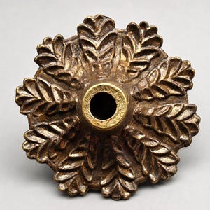 May include: A detailed, antique-style bronze-colored decorative furniture knob. The knob features a central hole surrounded by a ring of gold-colored metal. The outer edge is adorned with a repeating leaf pattern.