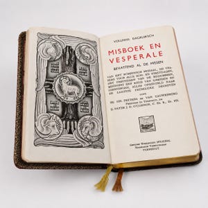 Puede incluir: Un libro abierto titulado MISBOEK EN VESPERALE con una ilustración en blanco y negro en la página izquierda que muestra un cordero central y cuatro figuras. La página derecha contiene texto en neerlandés en tinta negra y roja, con bordes dorados y dos marcapáginas.