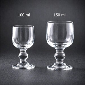 Op de afbeelding: Twee heldere wijnglazen met afgeronde kommen. Het kleinere glas is gemarkeerd met "100 ml" en het grotere met "150 ml". Beide glazen hebben een decoratieve, bolvormige steel en een ronde voet.