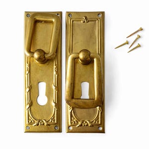 Op de afbeelding: Twee antieke messing deurklinkplaten met sleutelgaten en decoratieve bloemmotieven. Elke plaat heeft een rechthoekige vorm met een grote, rechthoekige handgreep. Drie kleine messing spijkers zijn ook zichtbaar.