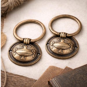 May include: Two antique brass drawer pulls with ring handles. Each pull features an oval backplate with a detailed urn design. The pulls are a warm, aged brass color, suggesting a vintage aesthetic. Ideal for furniture restoration or adding a classic touch.