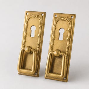 May include: A pair of antique brass door pulls. Each rectangular plate has a keyhole, an arched top, and a handle. The design incorporates floral and vine patterns. The pulls are a warm, aged gold colour.
