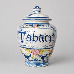 Puede incluir: Un tarro de cerámica con tapa, que presenta un esmalte de color crema y motivos florales pintados a mano en azul, amarillo y rosa. La palabra Tabac está escrita en una tipografía gótica azul oscuro sobre el cuerpo ancho del tarro.