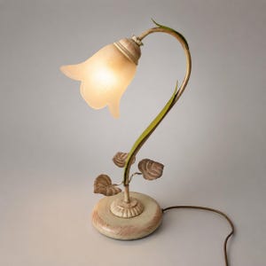 May include: A decorative table lamp with a floral design. The lamp features a cream-coloured, tulip-shaped shade and a curved stem with green and brown accents. The base is round and cream-coloured. The lamp is approximately 30 cm tall.
