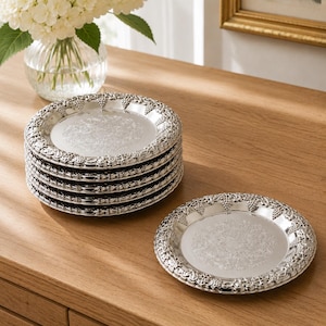 May include: A set of six round silver-toned plates featuring a reflective centre and a decorative border with a raised grape and vine motif. The plates have a polished metallic finish and are arranged in a stack with one plate placed separately.