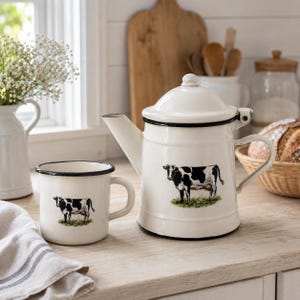 Könnte beinhalten: Eine weiße Emaille-Kaffeekanne und ein passender Becher, beide mit einer Illustration einer schwarz-weißen Kuh auf grünem Gras. Die Kaffeekanne hat einen Deckel und einen seitlichen Griff, während der Becher einen schwarzen Rand und einen Henkel hat.