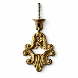 May include: An ornate brass drawer pull with a decorative, symmetrical design. The pull features a central floral motif and scrolled edges. A threaded screw is attached to the top for installation. The hardware has a vintage aesthetic.
