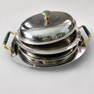 Peut inclure: Un ensemble de plats de service argentés avec un couvercle, un bouton turquoise et des poignées vertes et dorées. L'ensemble comprend un grand plat ovale et plusieurs plats imbriqués, reflétant l'environnement.