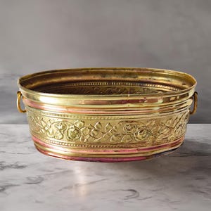 Op de afbeelding: Ovale messing plantenbak met reliëf bloemenontwerp. De plantenbak heeft een gouden afwerking met een roze band en twee kleine handvatten. Het bloemmotief omvat rozen en bladeren. De plantenbak is ongeveer 30 cm lang.