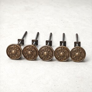 Op de afbeelding: Vijf antieke messing kastknoppen met sierlijke bloemmotieven. Elke knop heeft een ronde voorkant met een gedetailleerd patroon en een metalen steel voor montage. De knoppen zijn in een rij gerangschikt tegen een neutrale achtergrond.