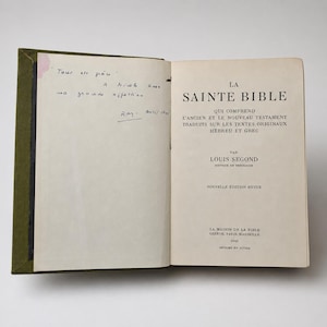 May include: An open book with a green cover and aged pages. The left page has handwritten notes in black ink, while the right page displays the title "LA SAINTE BIBLE" in bold black letters. The book is a vintage edition.