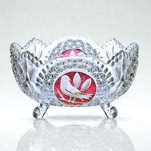 May include: A clear, cut-glass bowl with a scalloped rim and three small feet. The bowl features a red and white bird and leaf design. The bowl is likely used for serving or display.