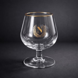 Op de afbeelding: Een helder cognacglas met een gouden rand en een gouden lauwerkransontwerp met de letter "N". Het glas heeft een brede kom, een korte steel en een ronde voet. Het glas staat tegen een donkere achtergrond.