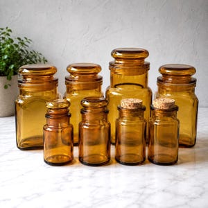 May include: A collection of eight amber-colored glass jars of varying sizes. Four larger jars feature matching amber glass lids, while four smaller jars are topped with natural cork stoppers. The jars have a square base and rounded shoulders.