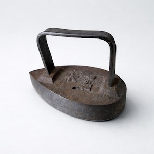 Puede incluir: Una plancha plana de hierro fundido, de color oscuro, con un asa curvada. La plancha tiene una forma redondeada, en forma de barco, y un aspecto envejecido. La superficie superior muestra marcas en relieve y un pequeño agujero.