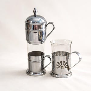 May include: A vintage chrome and glass coffee maker with a matching glass mug. The coffee maker has a cylindrical glass body with chrome accents, a lid, and a handle. The mug has a chrome holder with a sunburst design.