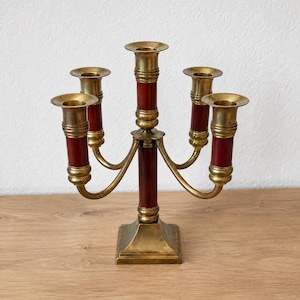 May include: A five-arm candelabra featuring a central pillar and four curved arms, all constructed from polished brass and deep red cylindrical accents. The piece stands on a square brass base with five circular candle holders at the top.